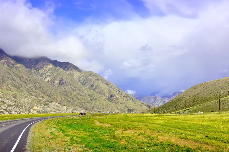A road through a green valley below cloud-topped mountains
