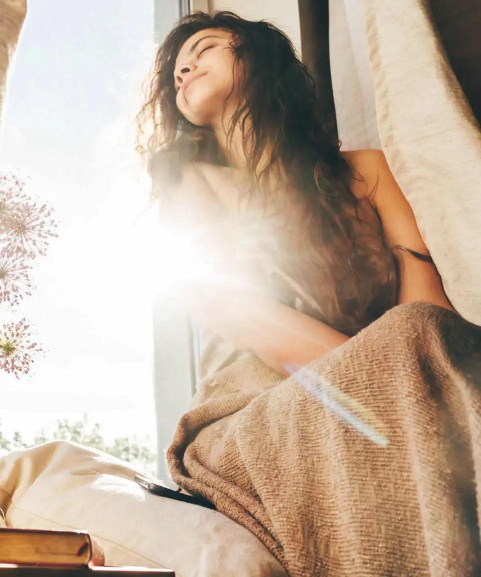 A woman relaxing by a sunlit window wrapped in a blanket